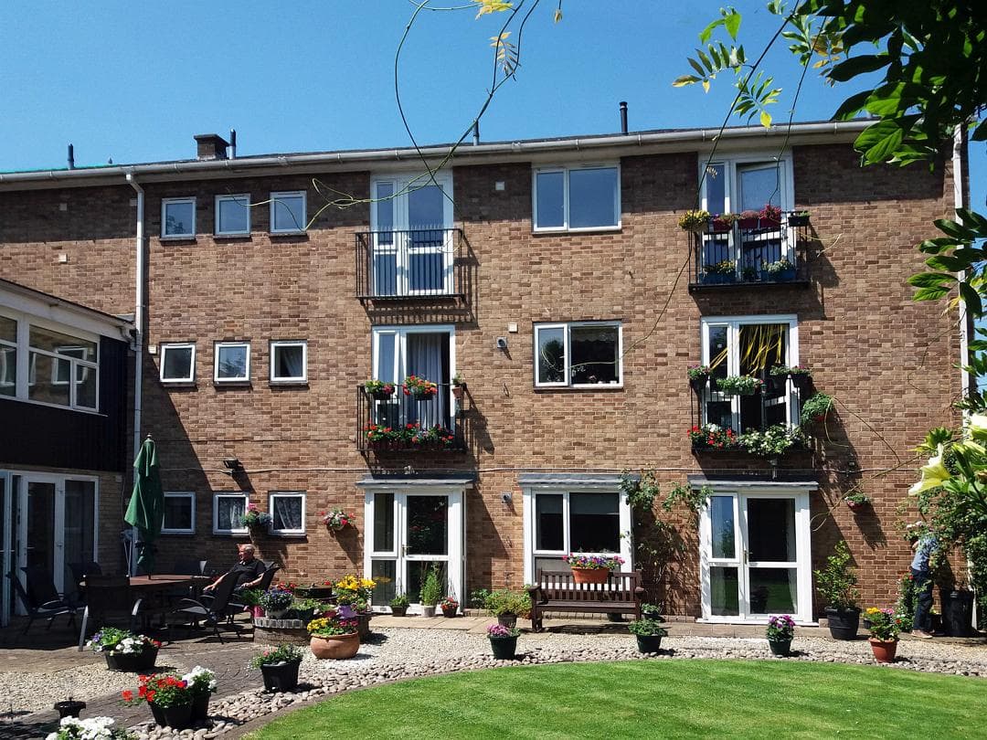 The garden front of the Colfe & Hatcliffe Glebe almshouses, a three-storey brick building with planted balconies, pots in flower and a lawn.