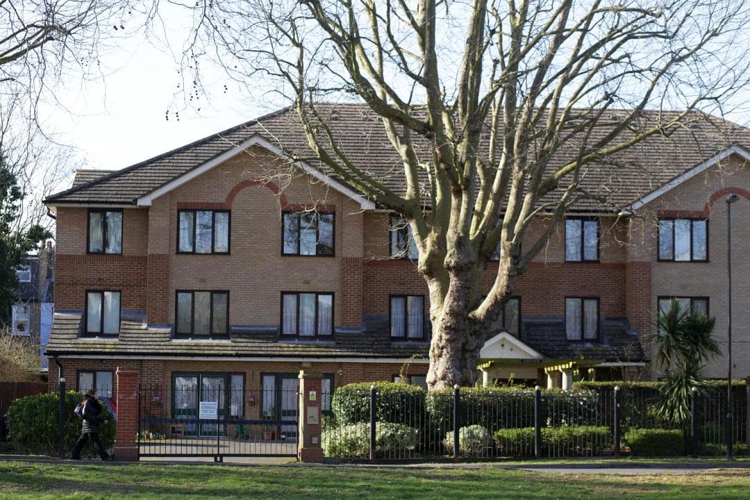 Lloyd Court almshouses from the green, a two-storey brick building behind railings with a large plane tree in front.