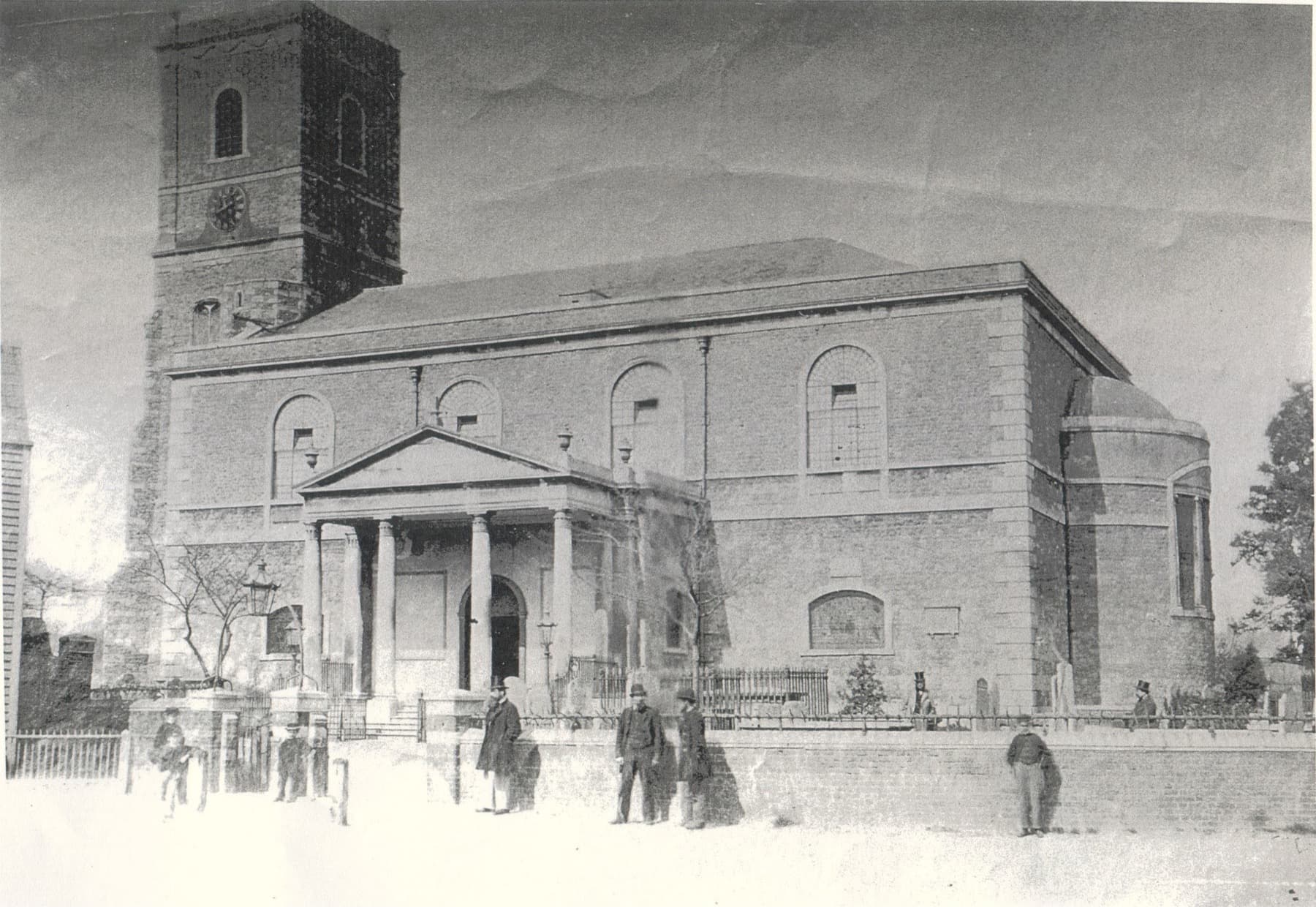 A Victorian photograph of the church: a tower with a clock, a pillared portico, and a rounded apse at the east end. Men in top hats stand by the churchyard railings.