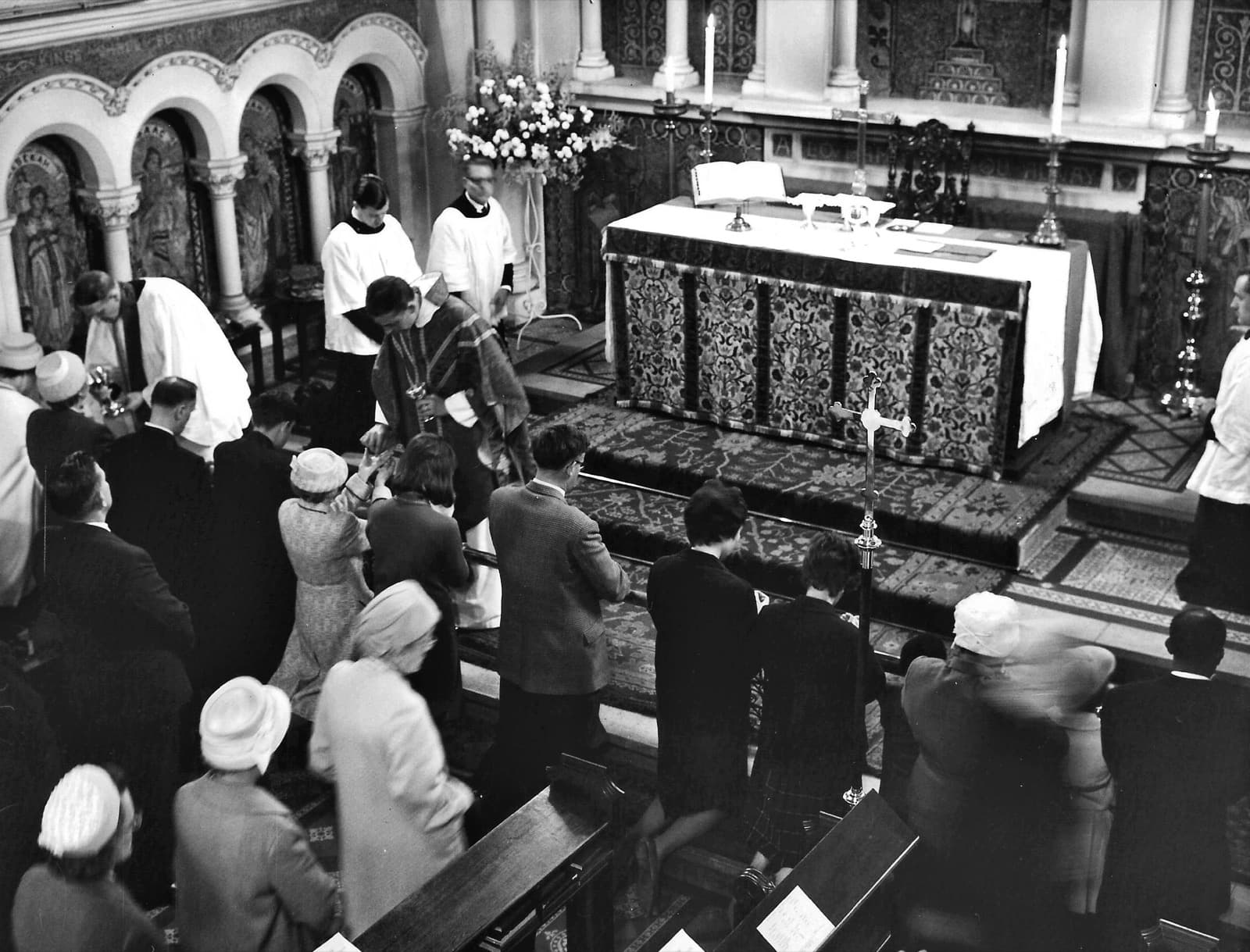 A row of people kneeling at an altar rail in the 1960s, most of the women in hats or headscarves, receiving communion from clergy in vestments.