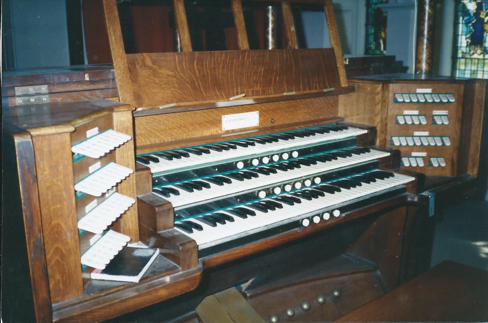 The same organ console in colour: light oak, three manuals, stop tabs in stepped rows on angled jambs either side, with the maker's nameplate above the top keyboard.