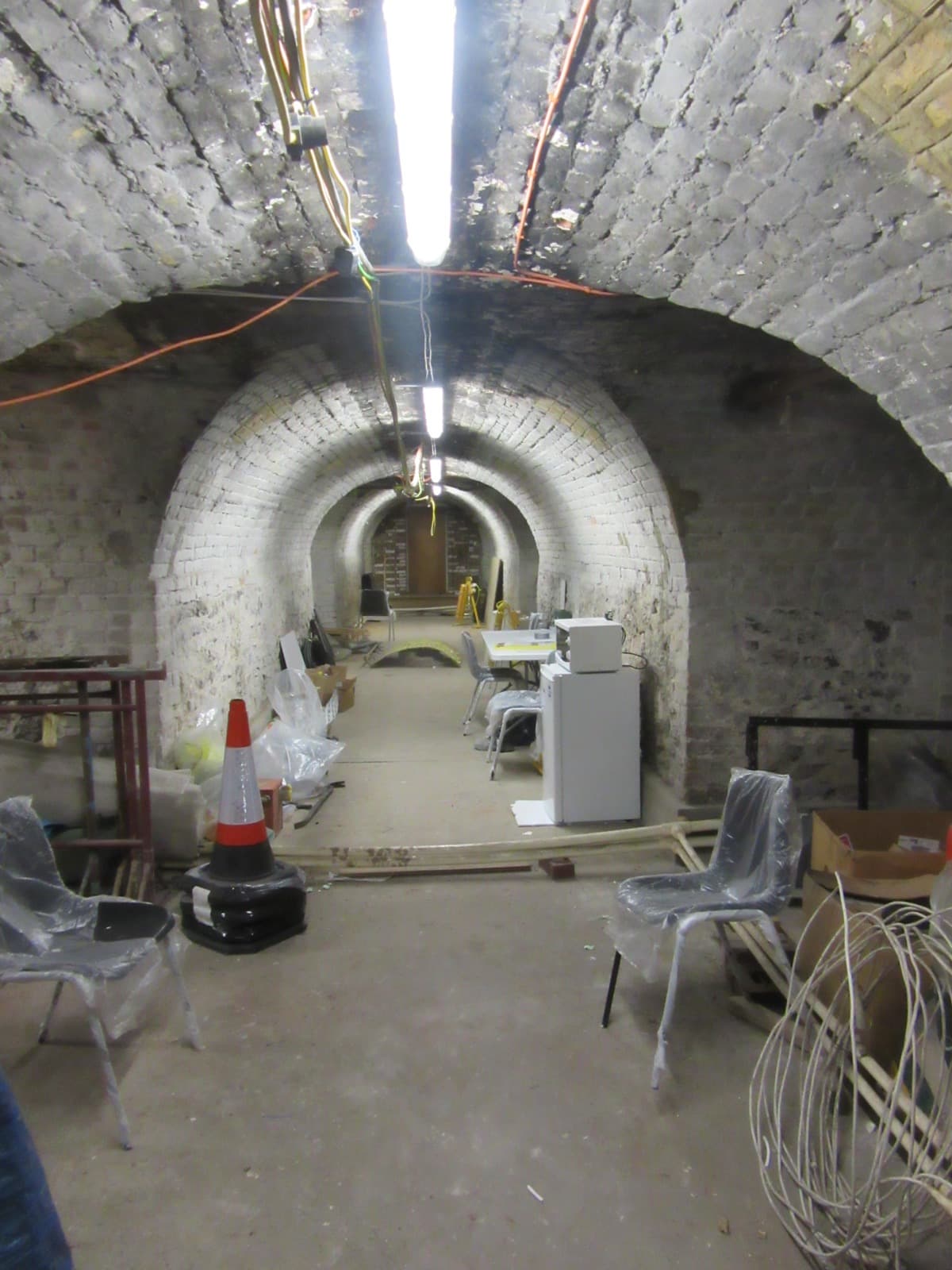 A whitewashed brick barrel-vaulted passage under the church, lit by a strip light, with chairs and equipment stored along it.