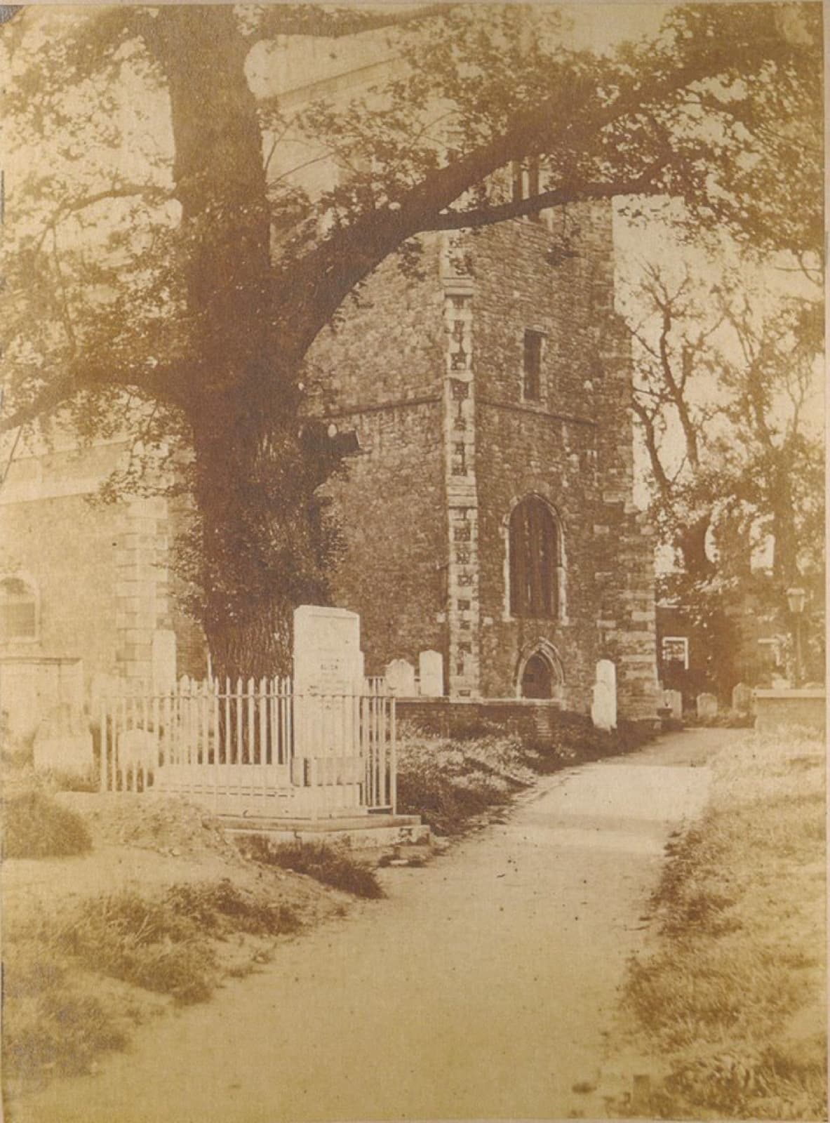 A sepia photograph of the church tower seen down a path, framed by the branches of a large tree, with a picket fence and gravestones.