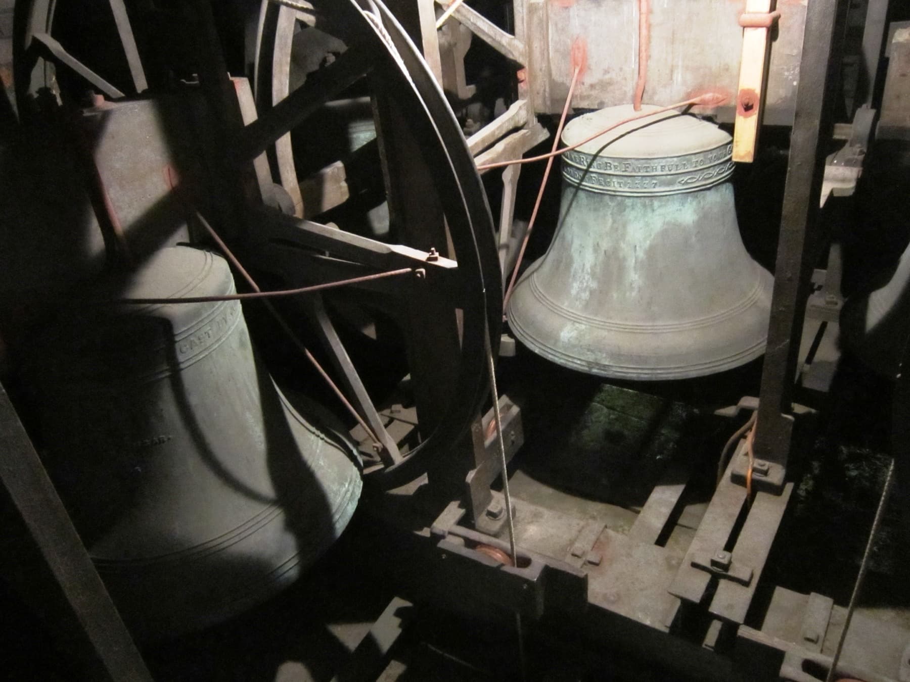A single bell hanging in the frame, seen close, with its wheel and headstock and the tower louvres behind.