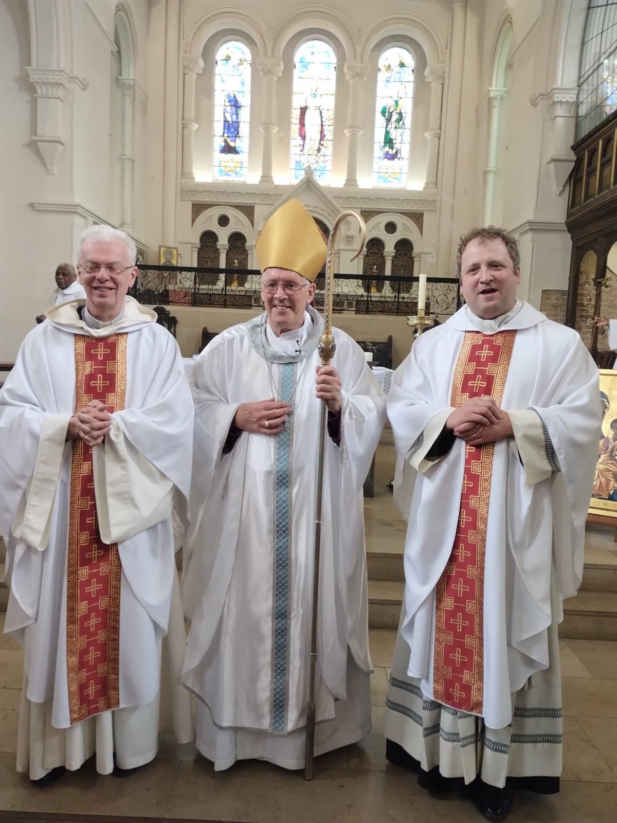 Three clergy in white vestments standing before the chancel. On the left, Fr Jürgen Francois, priest of St Peter's, Ghent; in the centre the Bishop of Southwark, in mitre and holding his crozier; on the right the Vicar of Lewisham. The icon of Our Lady of Lewisham is just visible at the right edge of the frame.