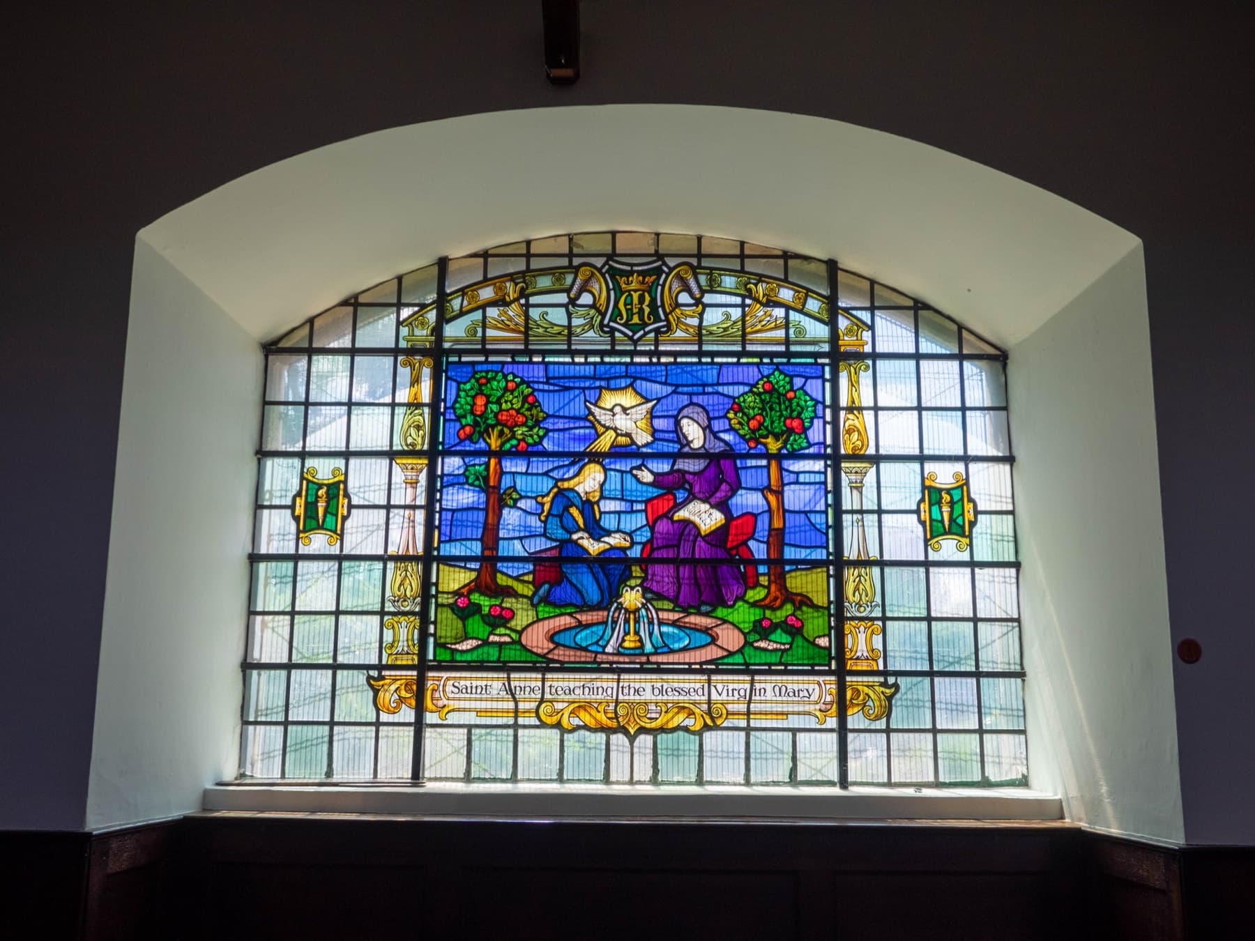 A stained glass window lit from outside, set in a plain arched recess.