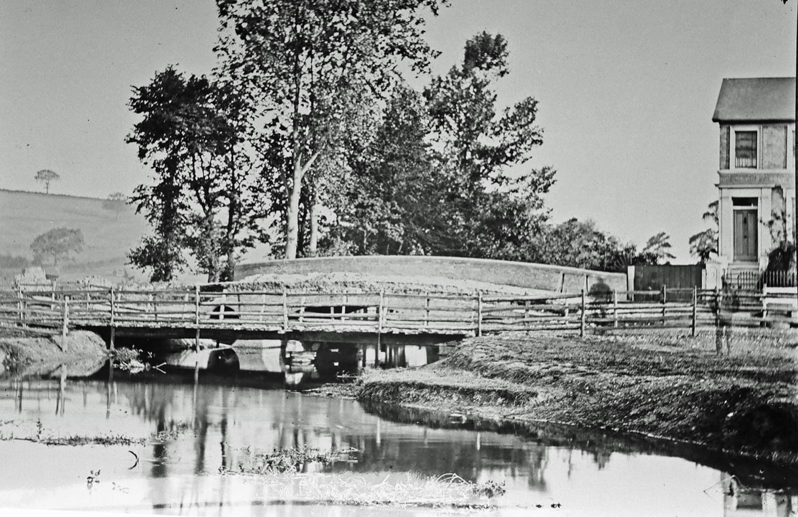 An old photograph of a wooden footbridge over a river, with houses beyond and trees on the bank.