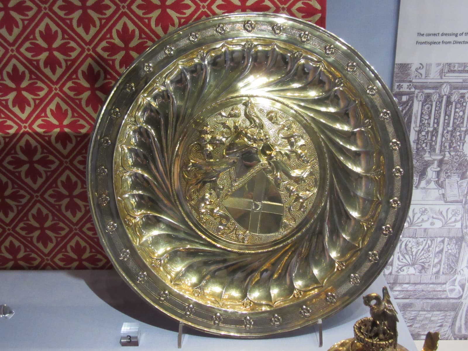 A large silver-gilt dish with swirling fluted decoration and an engraved centre, on display in a museum case.