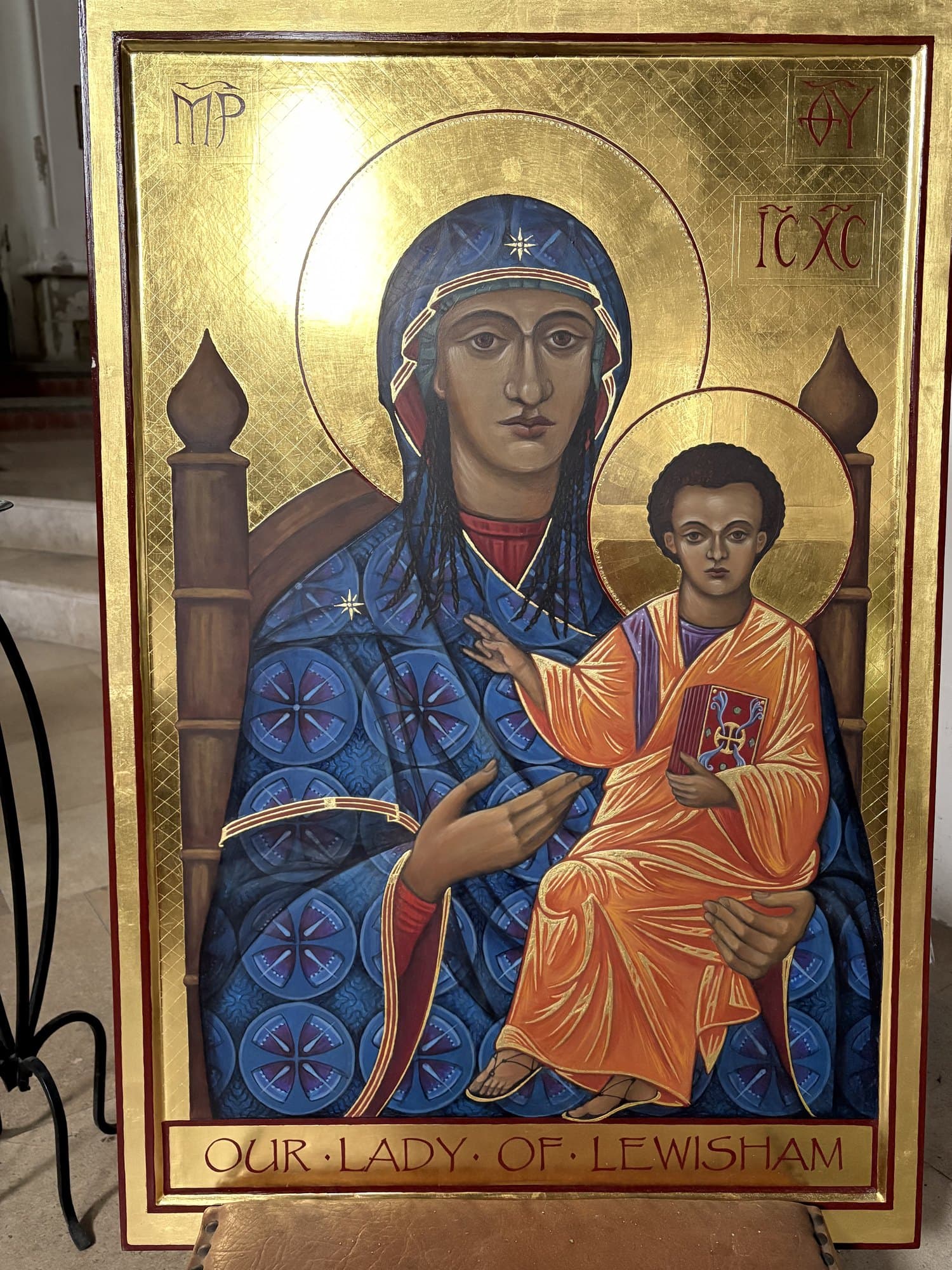 An icon of the Mother of God enthroned with the Christ Child on her lap, both with brown skin — she in a deep blue mantle patterned with rosettes in roundels, he in orange and gold holding a red book. The background is gold. Along the foot, in red capitals, OUR LADY OF LEWISHAM.