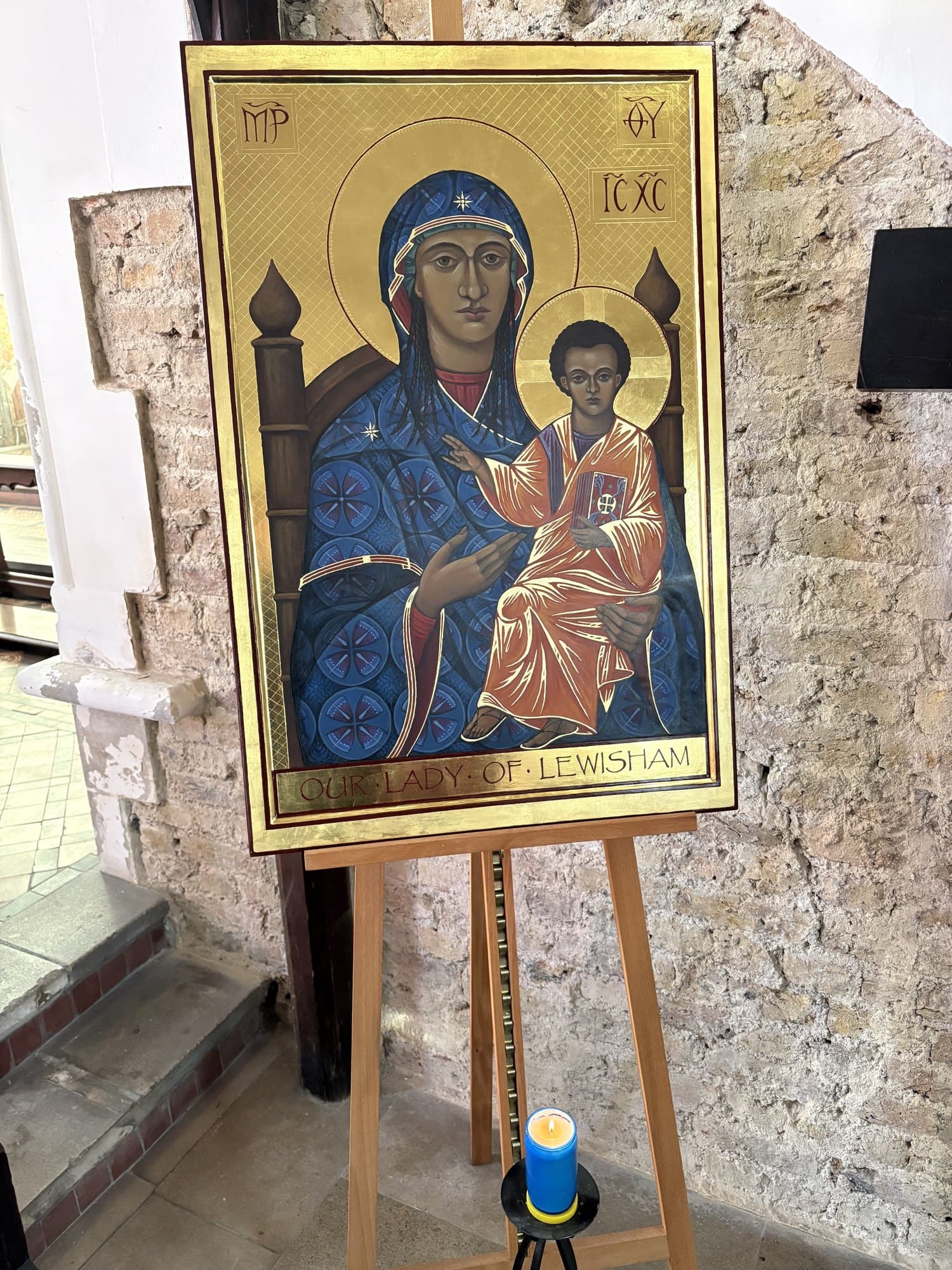 The same icon standing against the rough medieval stone of the church wall, with a blue votive candle burning on a stand in front of it.